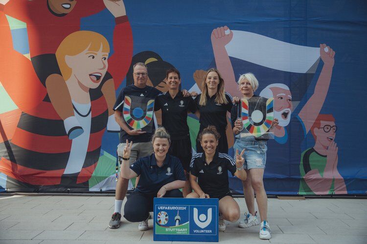 Ein Gruppen-Bild von 6 Helfer:innen vor einer bunten Wand. Sie tragen alle das gleiche blaue Hemd und lachen freundlich in die Kamera Zwei Personen halten Schilder von der UEFA EURO 2024 in der Hand.