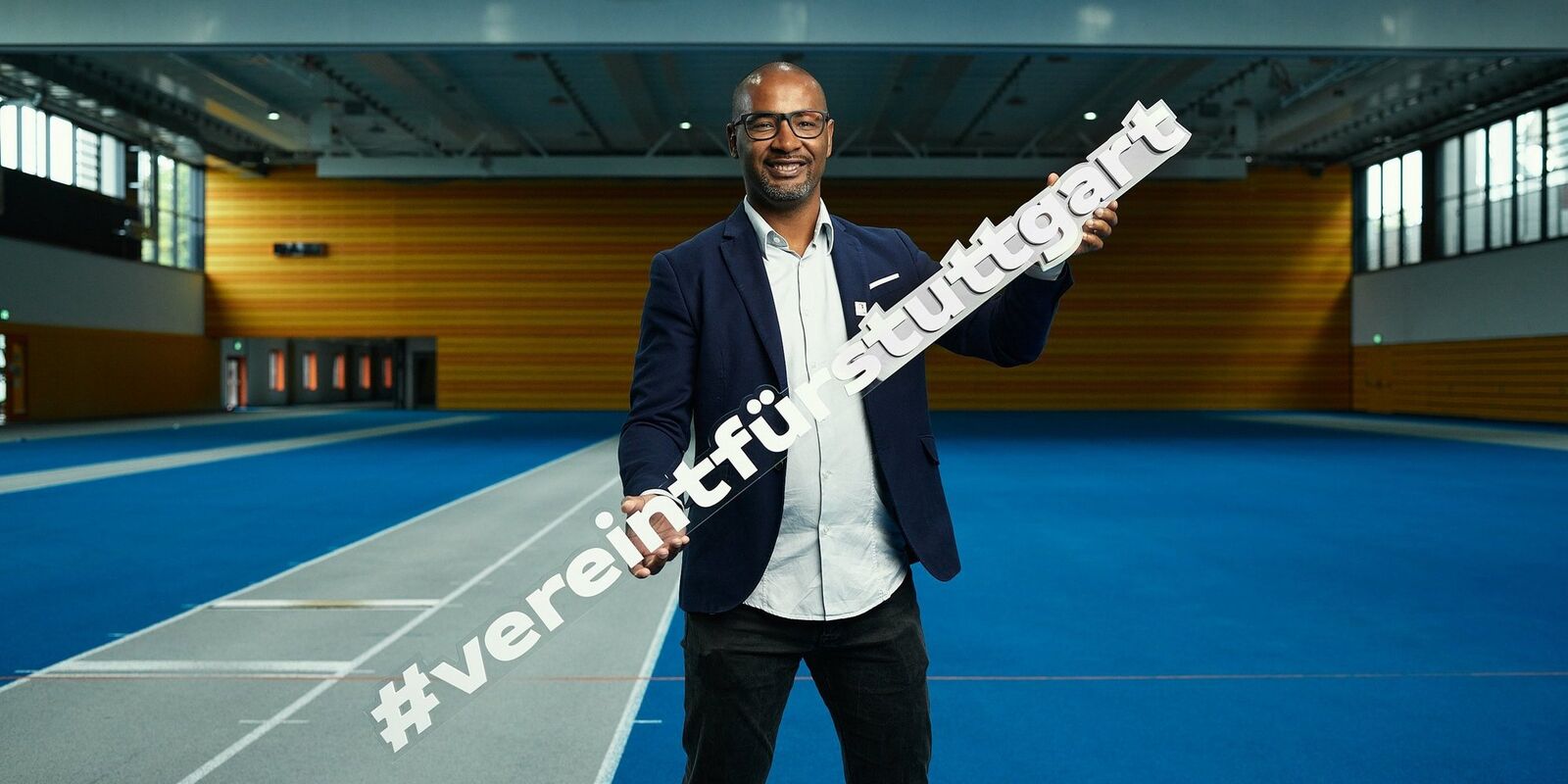 Foto von Cacau in einer Sport-Halle. Er trägt ein weißes Hemd und eine blaue feine Jacke. In den Händen hält er ein Schild mit den Worten: #vereintfürstuttgart. 