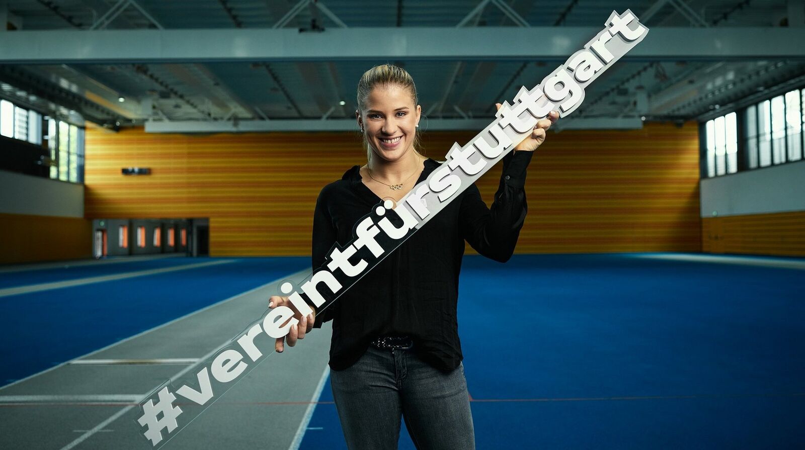 Foto von Elisabeth Seitz in einer Sport-Halle. Sie ist blond und hat die Haare zusammengebunden. Sie trägt einen schwarzen Pulli und eine graue Hose.In den Händen hält sie ein Schild mit den Worten: #vereintfürstuttgart. 