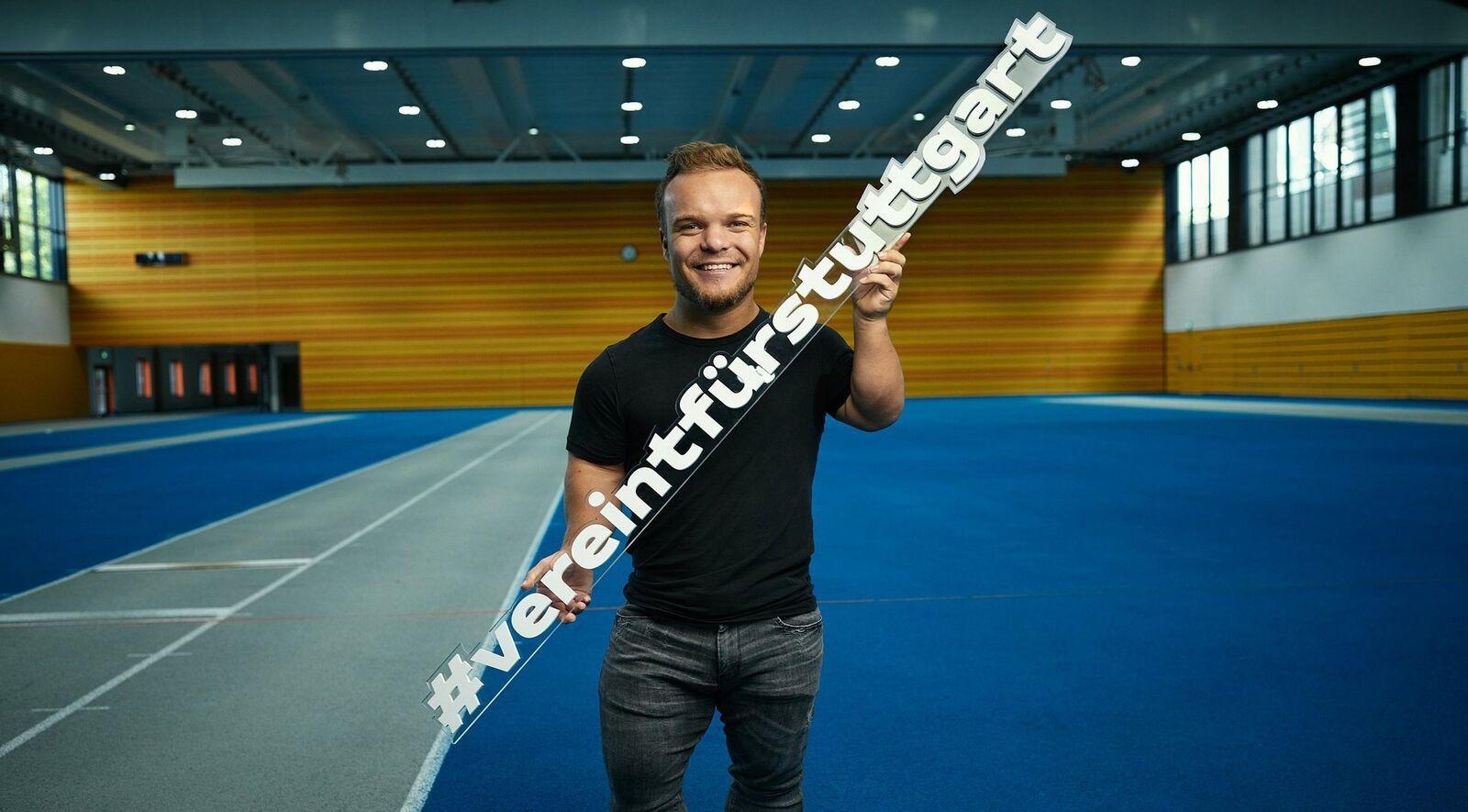 Foto von Niko Kappel in einer Sport-Halle. Er hat blonde Haare und einen Bart. Er trägt ein schwarzes T-Shirt und eine graue Hose.In den Händen hält er ein Schild mit den Worten: #vereintfürstuttgart. 